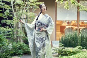 ผู้หญิงสวมชุดกิโมโนแบบดั้งเดิมยืนอยู่ในสวนญี่ปุ่นที่เงียบสงบ ล้อมรอบด้วยต้นไม้และอาคารสไตล์ญี่ปุ่นในเกียวโต